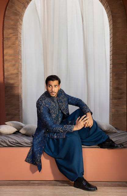 A man wearing a traditional Indian outfit consisting of a navy blue embroidered kurta and matching trousers, sitting on a terracotta-colored window seat with white curtains and brick arches in the background.