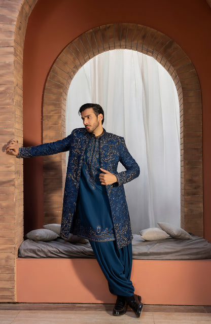 A man models a traditional Indian outfit consisting of a navy blue embroidered sherwani jacket over a matching navy kurta and trousers, posed in front of an arched brick window with white curtains.