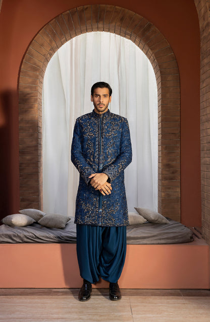 A man wearing a traditional Indian outfit consisting of a navy blue embroidered sherwani and matching trousers, standing in front of an arched brick opening with white curtains and a bed in the background.