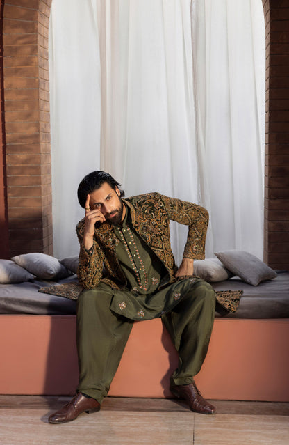 A man wearing an olive green and gold embroidered traditional outfit sits on a cushioned bench in front of white curtains, with brick columns on both sides.