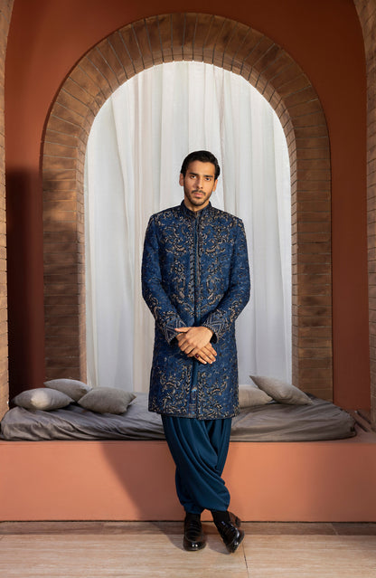 A man wearing a traditional Indian outfit stands in front of an arched brick opening with white curtains and a cushioned bench. The outfit consists of a long navy blue sherwani with intricate gold embroidery, paired with dark blue pants and black shoes.