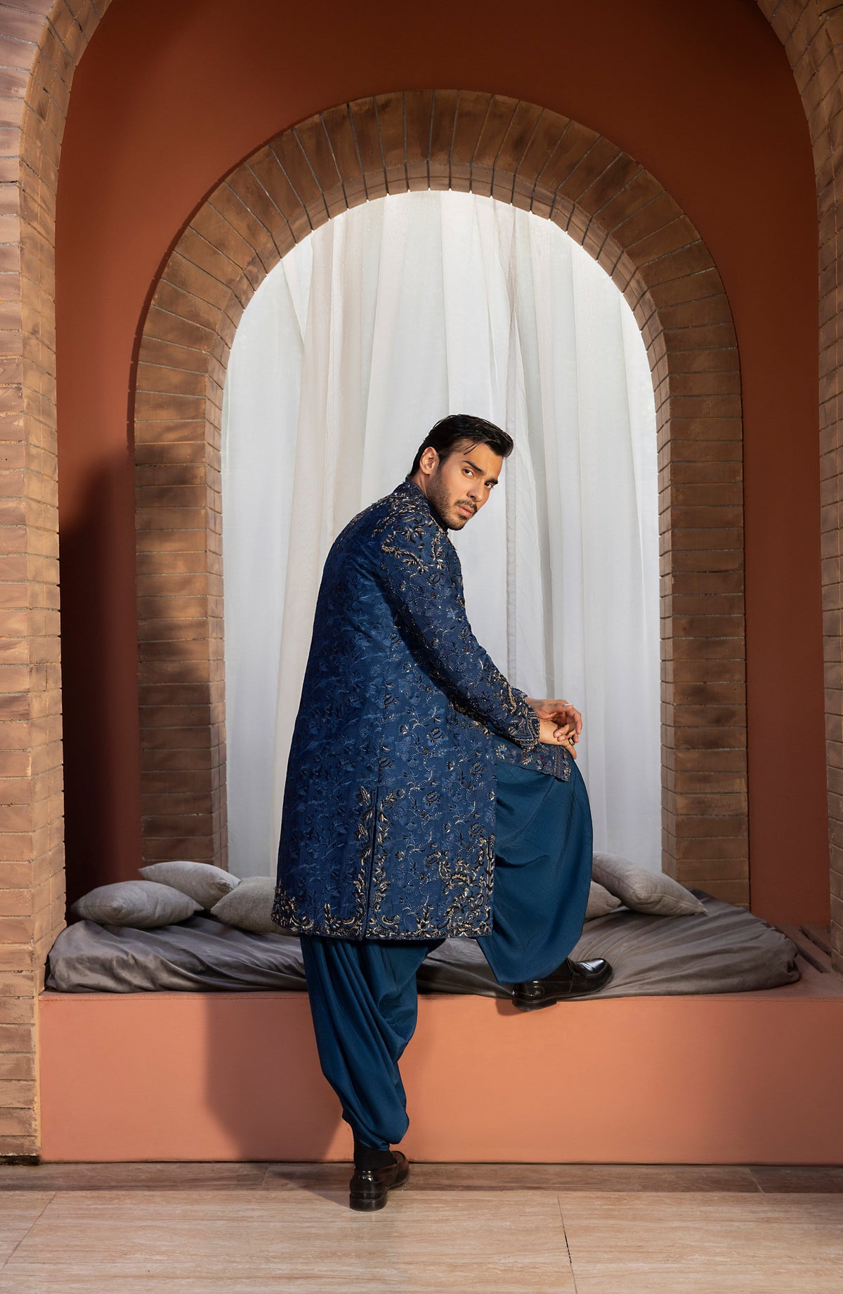A man wearing a traditional Indian outfit, featuring a navy blue embroidered kurta and matching trousers, posing in front of a brick archway with a bed in the background.