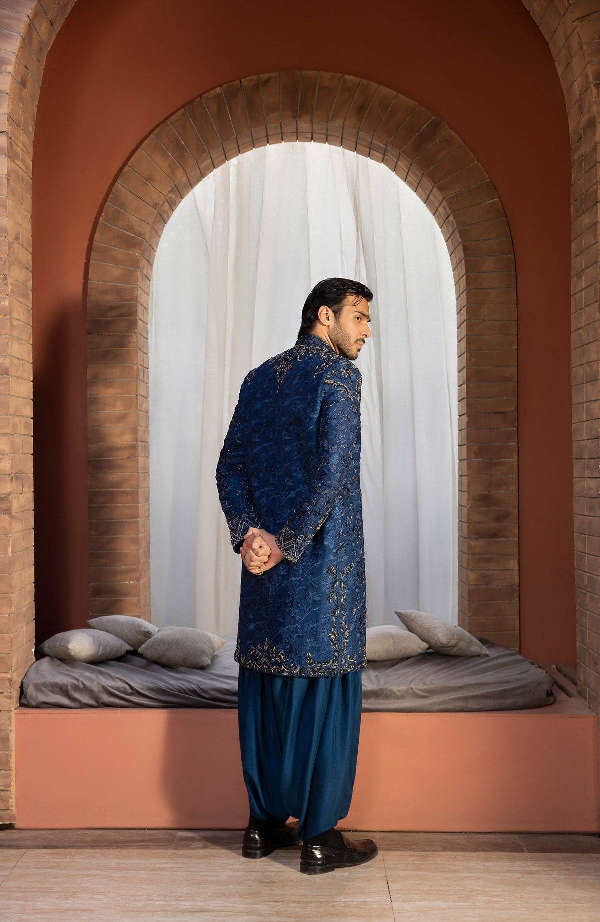 A man wearing a traditional blue embroidered sherwani and matching trousers stands in profile inside a terracotta-colored arched alcove with a built-in bed and white curtains behind him.