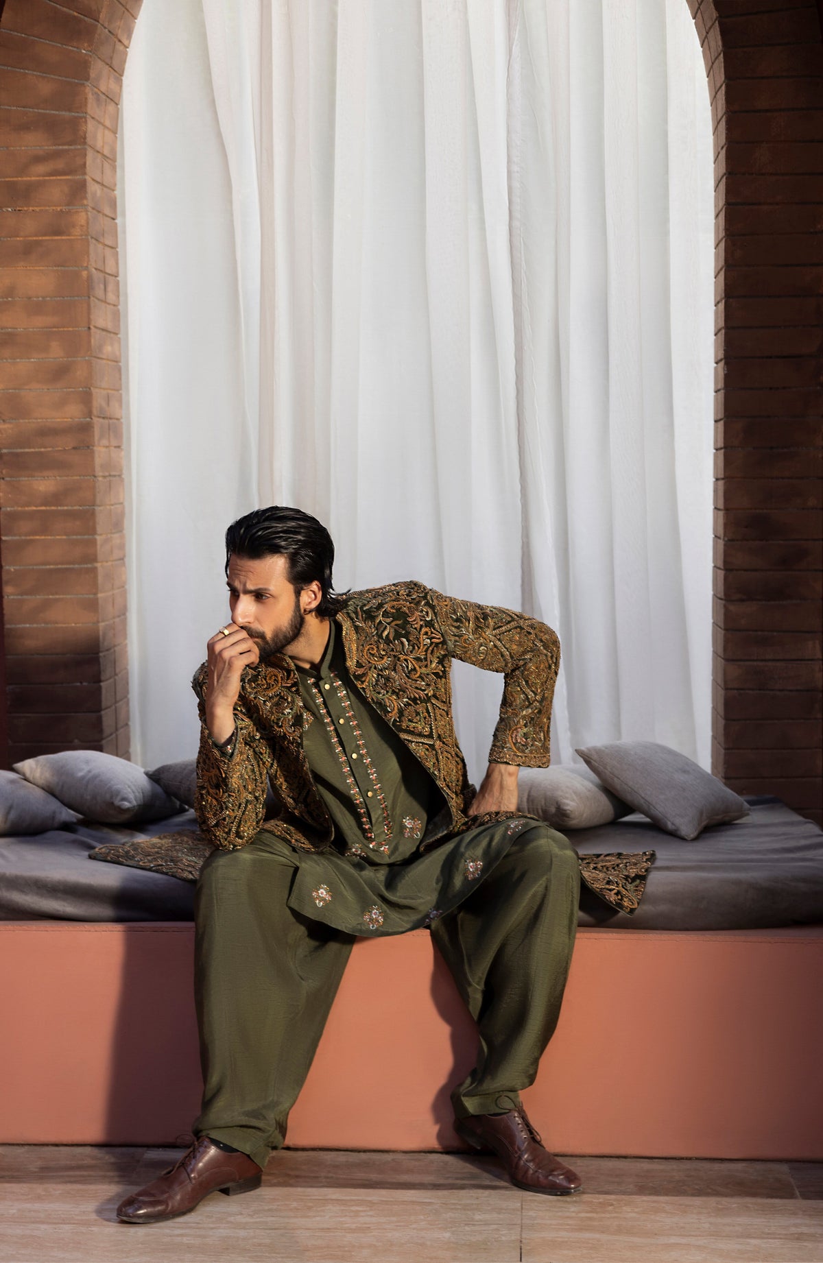 A man wearing an olive green and gold embroidered traditional outfit sits on a cushioned bench in front of white curtains and brick arches.