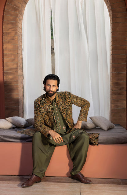 A man wearing an ornate olive green and gold embroidered traditional outfit sits on a cushioned bench in front of white curtains and brick arches.