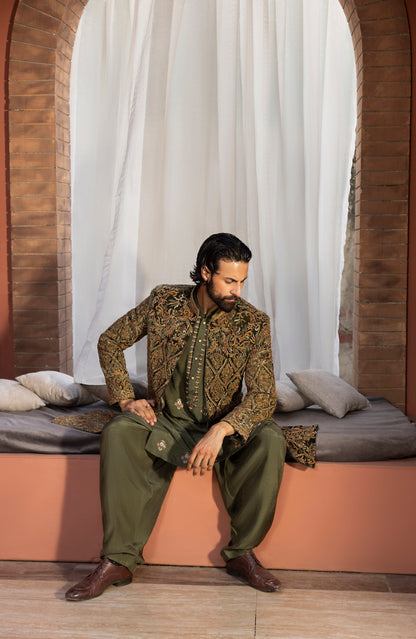 A man wearing an olive green and gold patterned traditional South Asian outfit sits on a peach-colored platform in front of white curtains and brick arches.