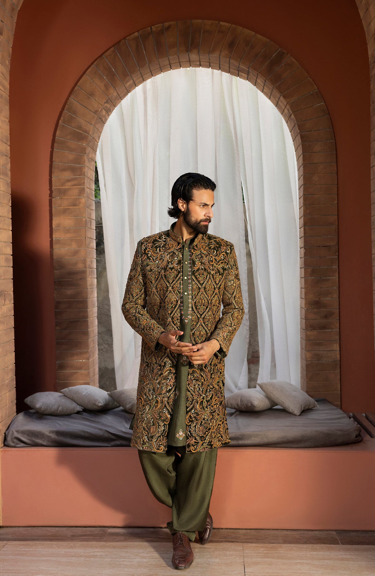 A man wearing a traditional South asian outfit with an olive green kurta featuring intricate gold and brown embroidery, paired with olive green trousers, standing in front of a rustic arched doorway with white drapes.