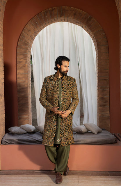 A man wearing a traditional South asian outfit with an olive green kurta featuring intricate gold and brown embroidery, paired with olive green trousers, standing in front of a rustic arched doorway with white drapes.