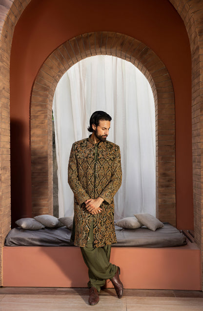 A man wearing a traditional South Asian outfit stands inside a brick archway alcove. The outfit consists of a brown and gold patterned sherwani jacket, olive green trousers, and brown leather shoes.