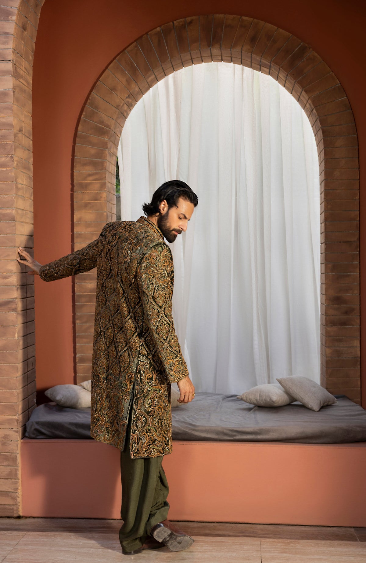 A man wearing a long-sleeved brown and gold patterned sherwani with green trousers stands in front of a large arched window with white curtains and a built-in bed.