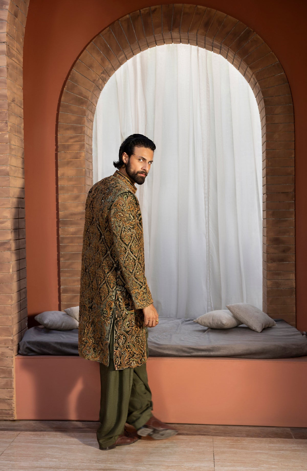 A man wearing a long-sleeved patterned brown and gold sherwani with green trousers stands in profile in front of a large arched window with white curtains and a built-in bed.