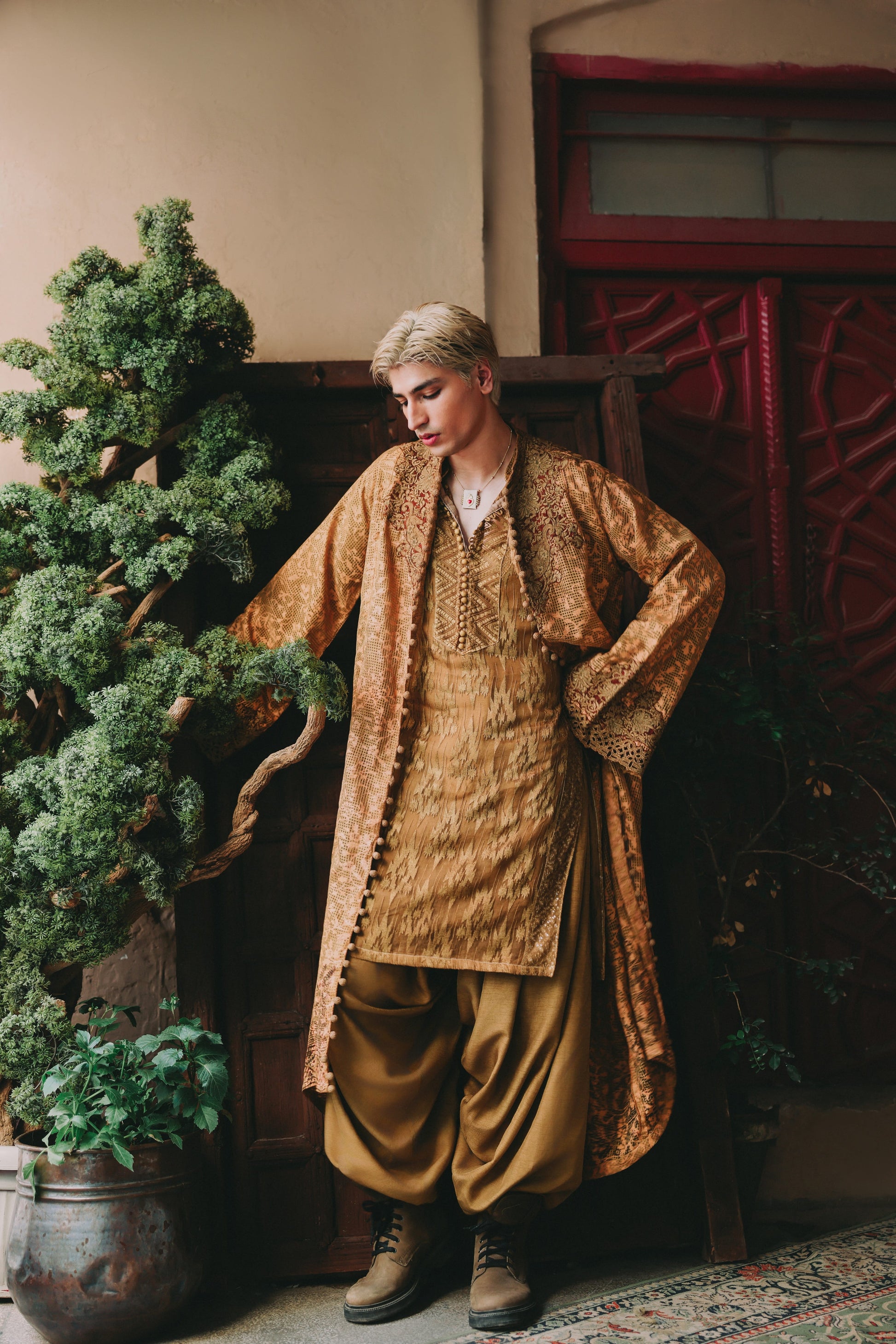 Person wearing a traditional outfit standing next to a decorative plant indoors.