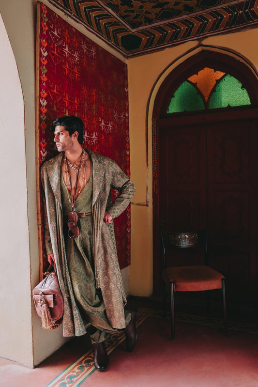 Man in traditional attire standing in a room with decorative walls and a chair.