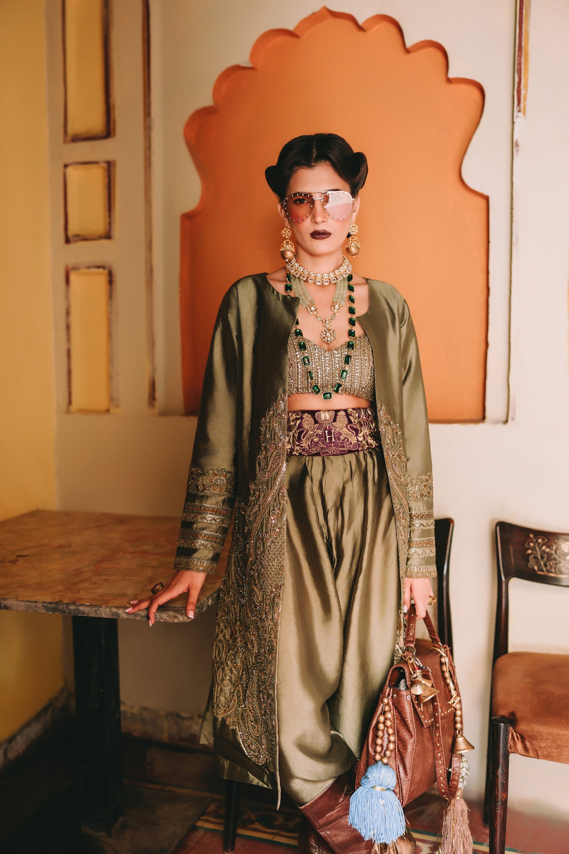 Woman in a green dress with intricate patterns and jewelry, holding a brown handbag, standing in a room with an orange wall and wooden furniture.