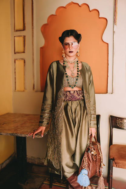 Woman in a green dress with intricate patterns and jewelry, holding a brown handbag, standing in a room with an orange wall and wooden furniture.