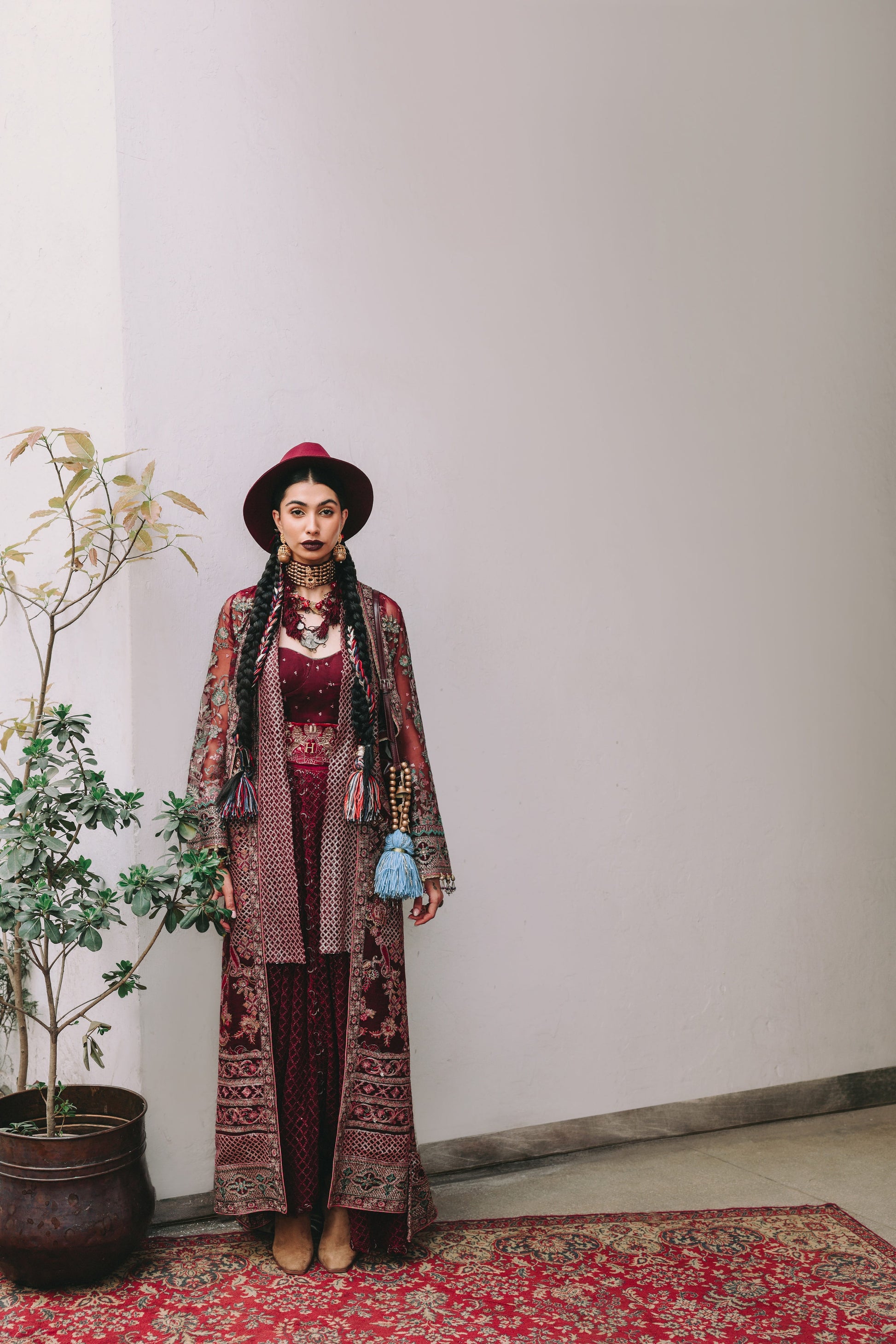 Woman in a patterned outfit standing against a plain wall with a plant to her left.