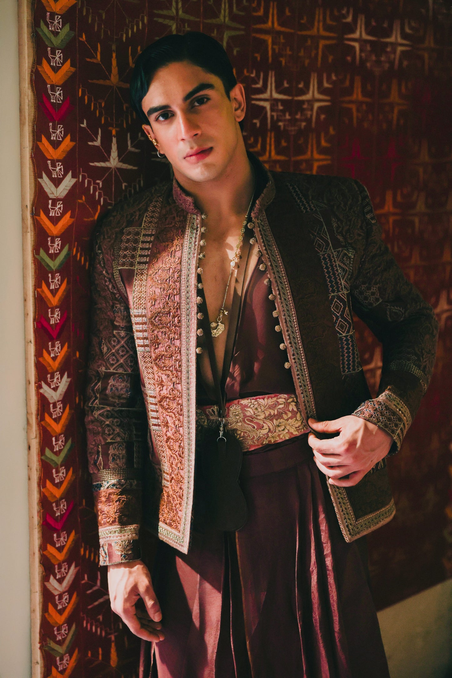 Man in ornate traditional outfit standing against a patterned wall.