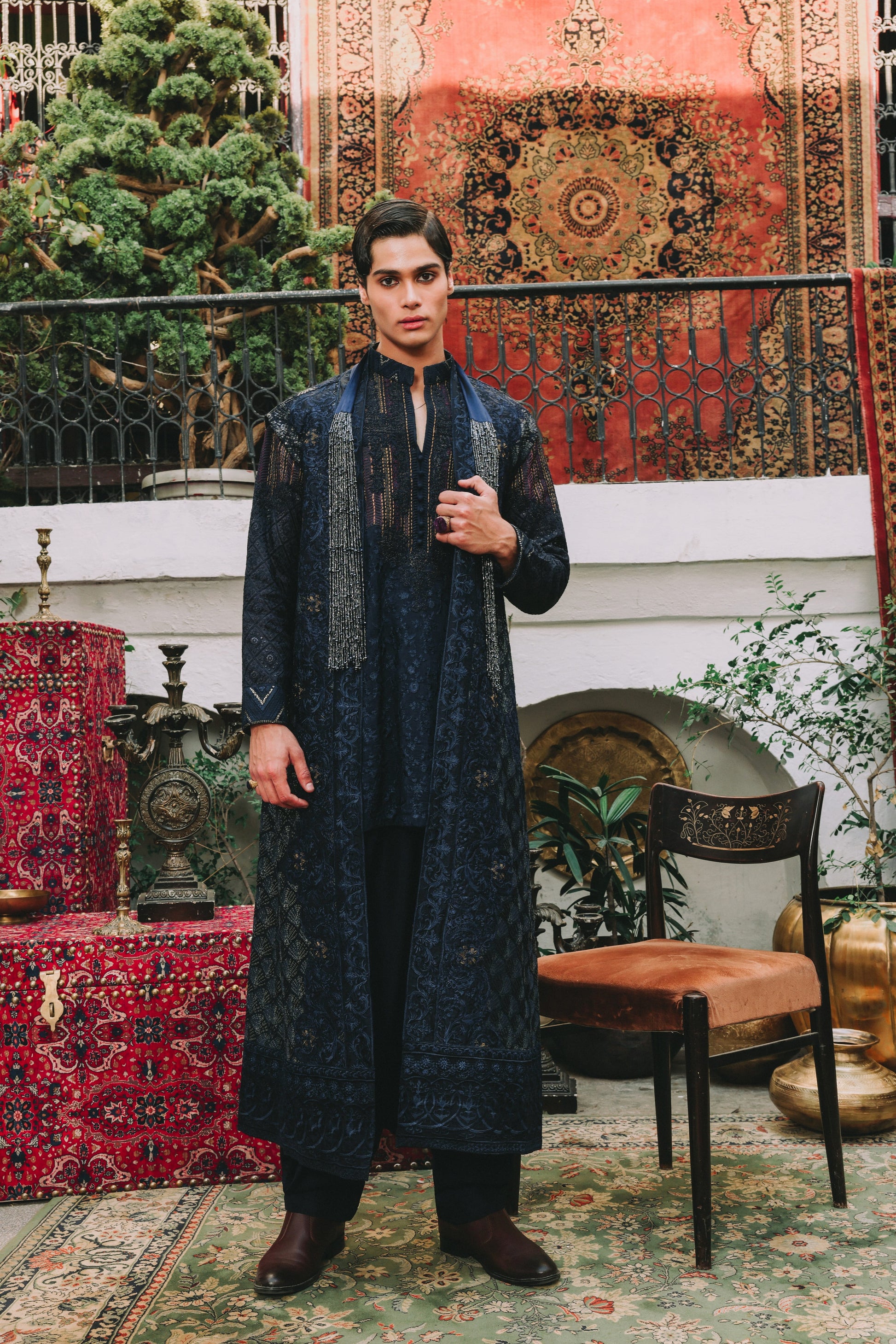 Man in a dark blue traditional outfit standing in a decorative room with patterned rugs and furniture.