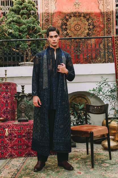 Man in a dark blue traditional outfit standing in a decorative room with patterned rugs and furniture.