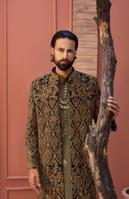 A man wearing a long-sleeved olive green sherwani jacket with intricate gold and brown embroidery, standing next to a large tree trunk in front of a terracotta-colored wall.