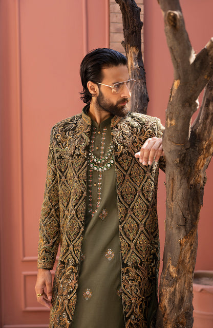 A man wearing a traditional South Asian outfit with an olive green kurta and a heavily embroidered gold and brown sherwani jacket, standing next to a tree trunk in front of a dusty pink wall.