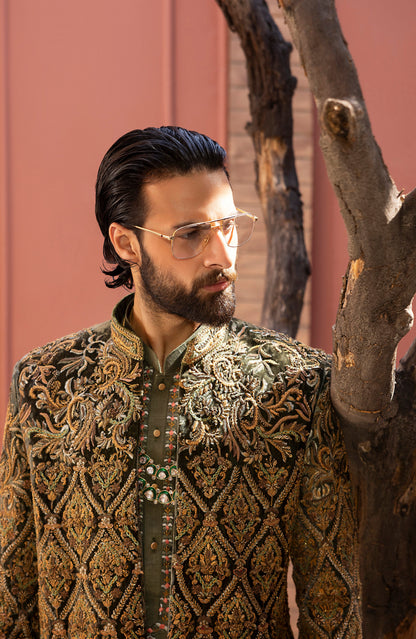 A man wearing a heavily embroidered olive green and gold sherwani jacket with gold-framed glasses, standing next to a tree trunk against a dusty rose wall.