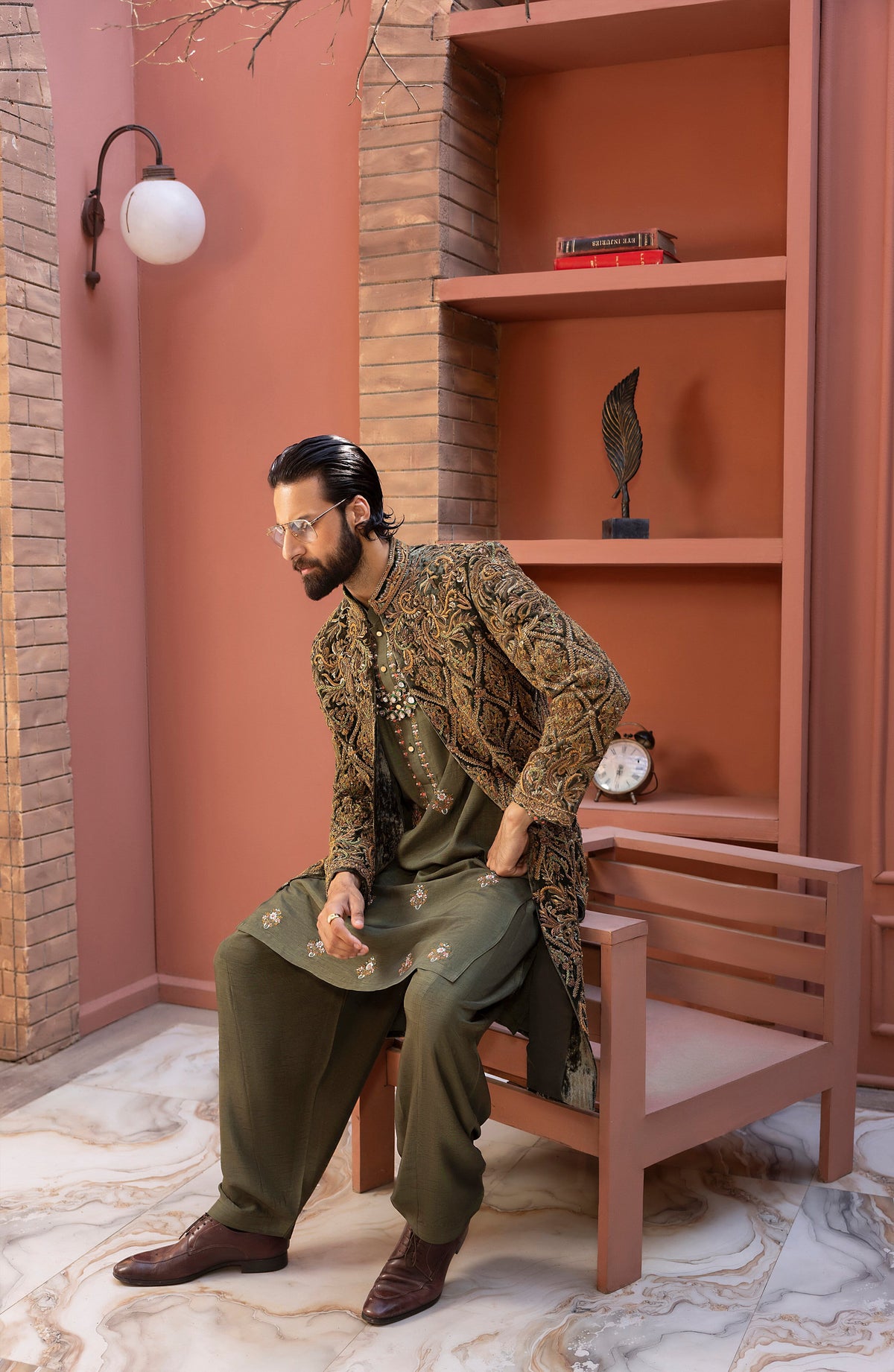 A man wearing a traditional South Asian outfit with a heavily embroidered olive green kurta and matching trousers sits on a wooden chair in a styled room with terracotta-colored walls and brick accents.