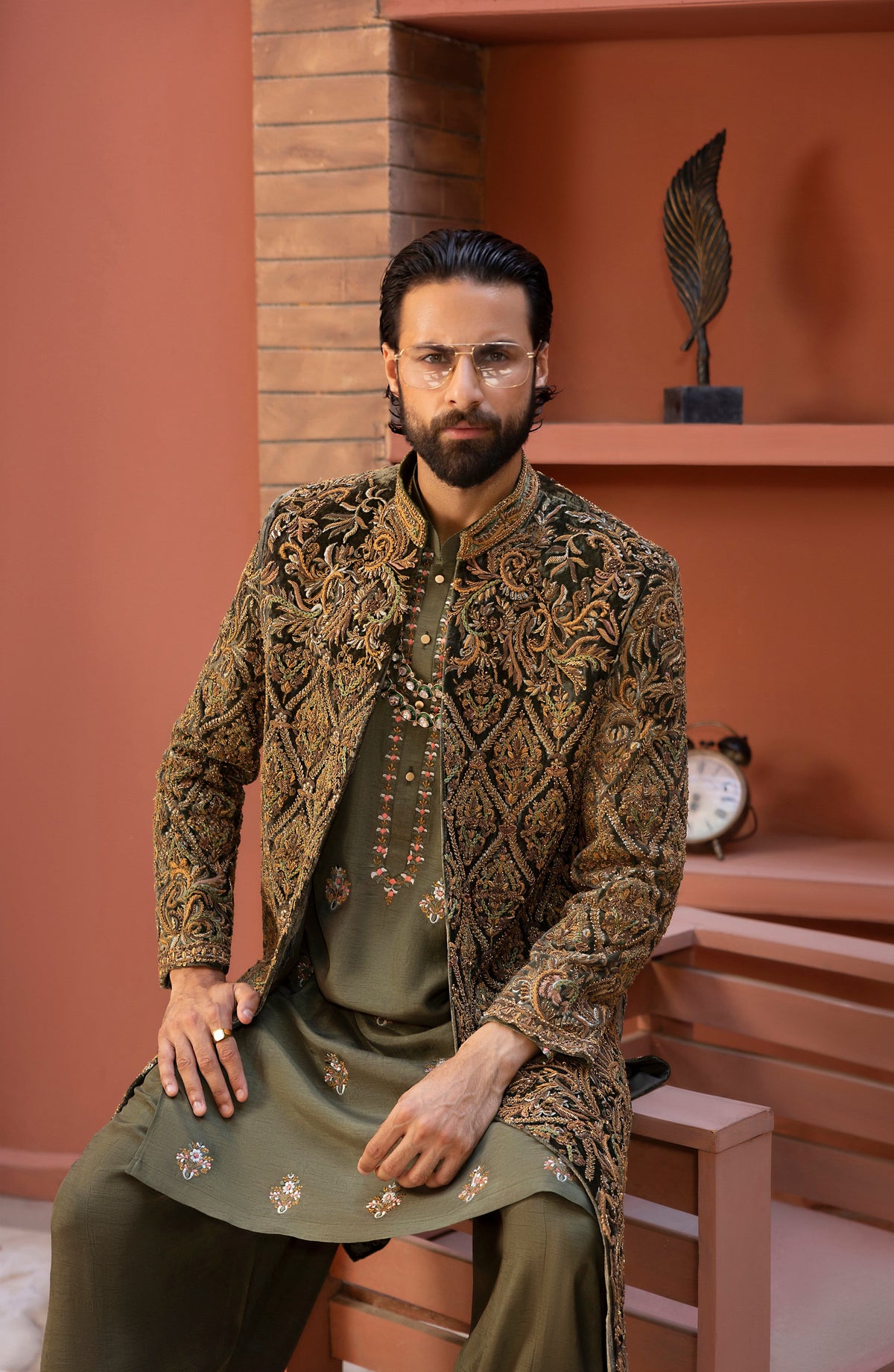 A man wearing a detailed gold and brown embroidered sherwani jacket over an olive green kurta and trousers, sitting on a wooden bench in a warm terracotta-colored room.