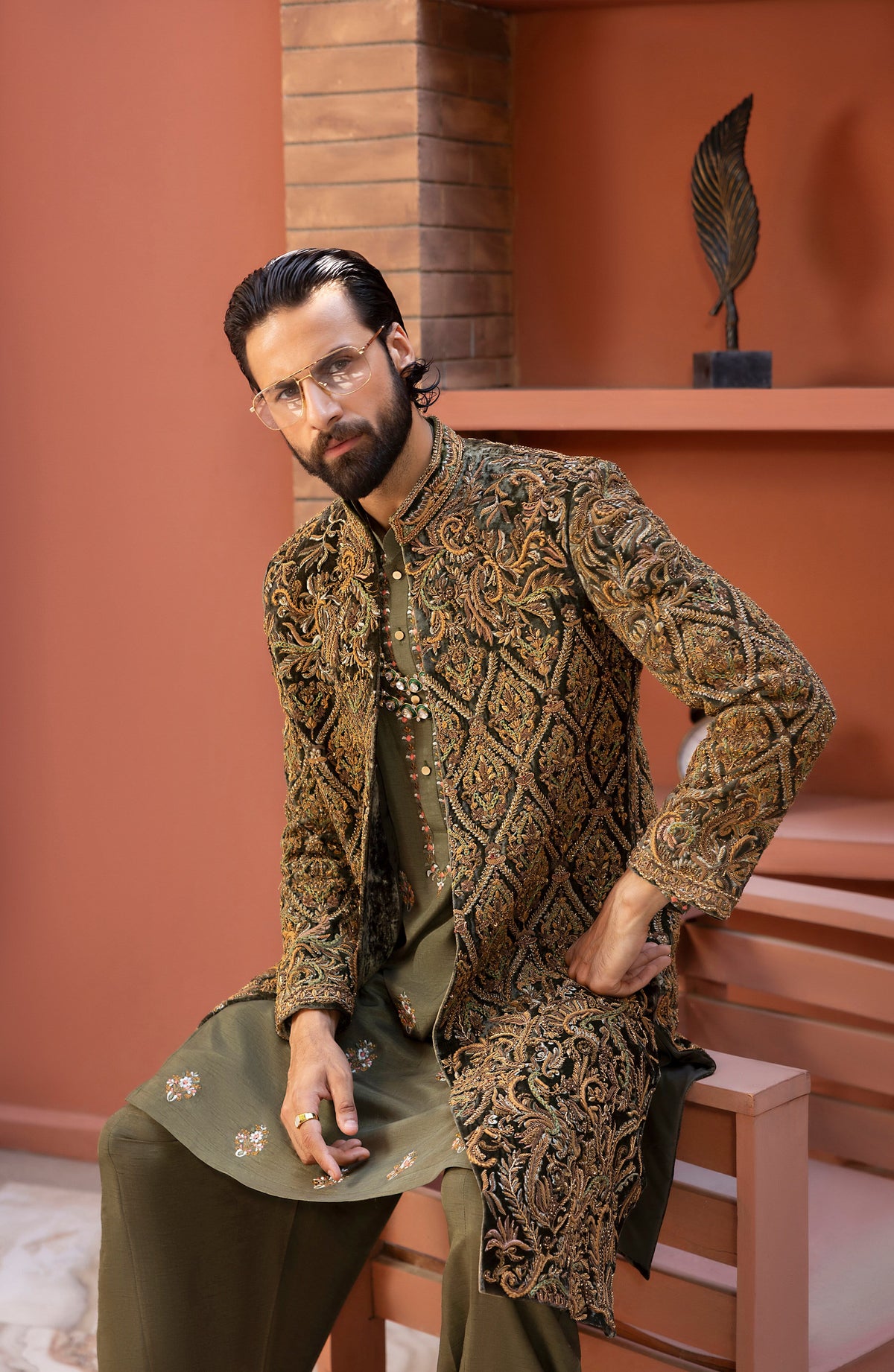 A man wearing a traditional South Asian outfit with an ornate gold and brown embroidered sherwani jacket over an olive green kurta and trousers, sitting on a wooden bench in a terracotta-colored room.