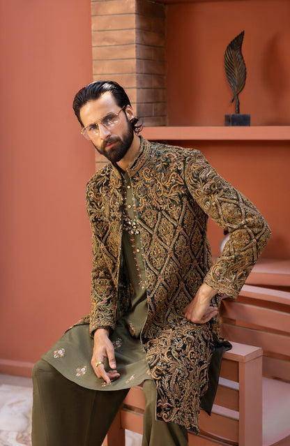 A man wearing a traditional South Asian outfit with an ornate gold and brown embroidered sherwani jacket over an olive green kurta and trousers, sitting on a wooden bench in a terracotta-colored room.