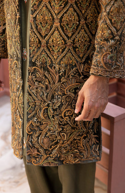 Close-up of a person wearing an ornate embroidered tunic with intricate gold, brown, and green paisley and floral patterns over a dark base, paired with olive green trousers.