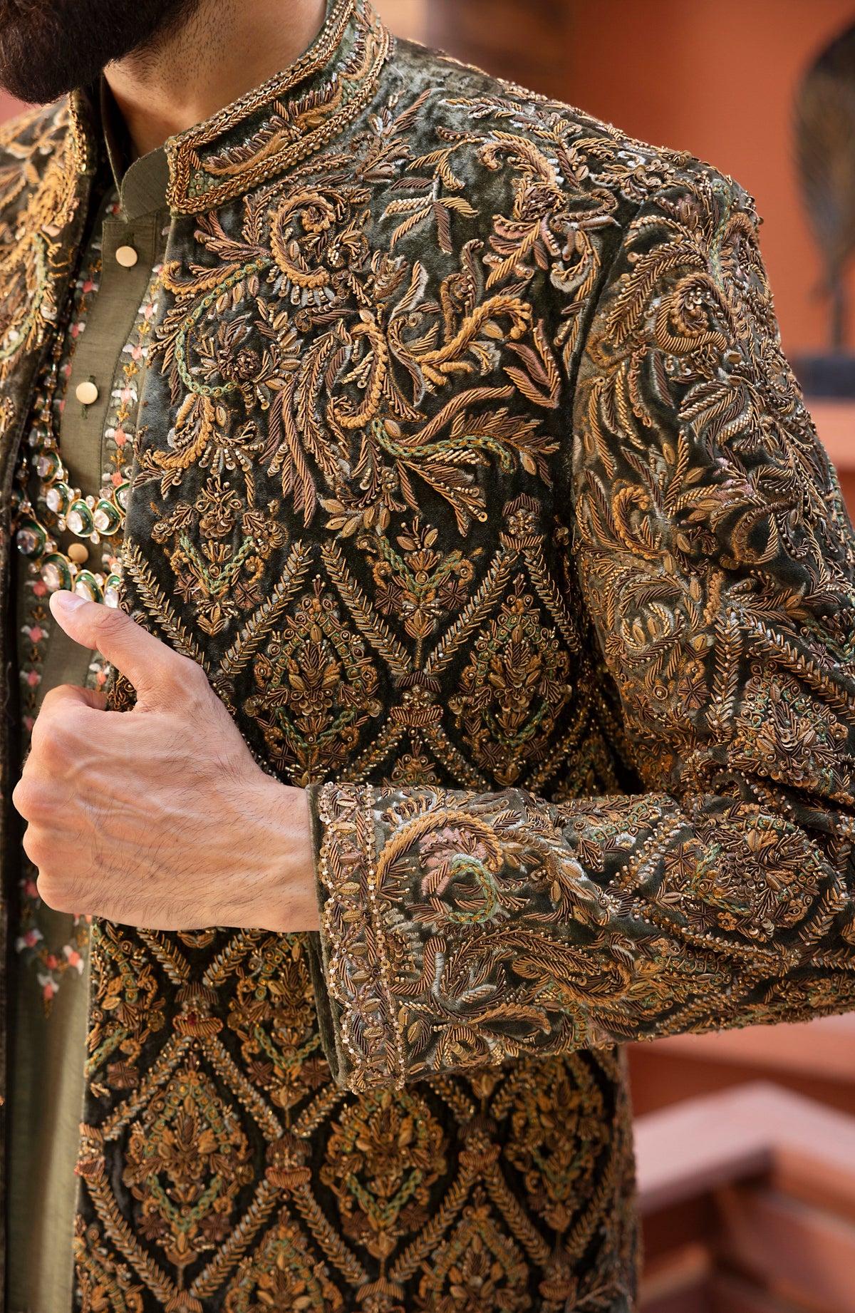 Close-up of a man wearing an ornate dark olive green sherwani jacket with intricate gold and brown embroidery, paired with a matching olive green shirt.