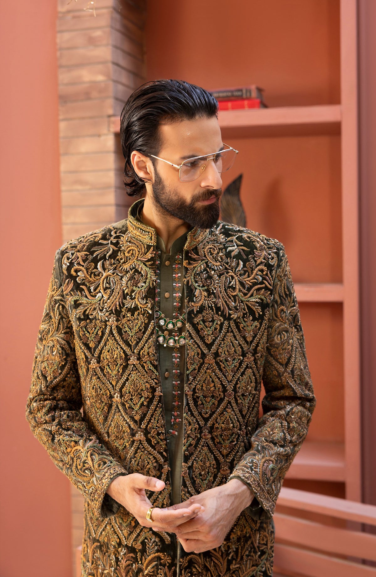 A man wearing a highly detailed, ornate gold and brown embroidered sherwani jacket with a mandarin collar, styled with a dark green inner shirt and thin metal-framed glasses, standing in front of a terracotta-colored brick wall and wooden shelves.
