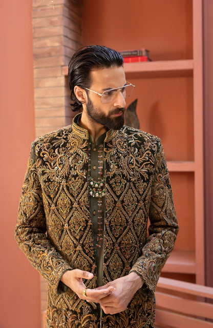 A man wearing a highly detailed, ornate gold and brown embroidered sherwani jacket with a mandarin collar, styled with a dark green inner shirt and thin metal-framed glasses, standing in front of a terracotta-colored brick wall and wooden shelves.