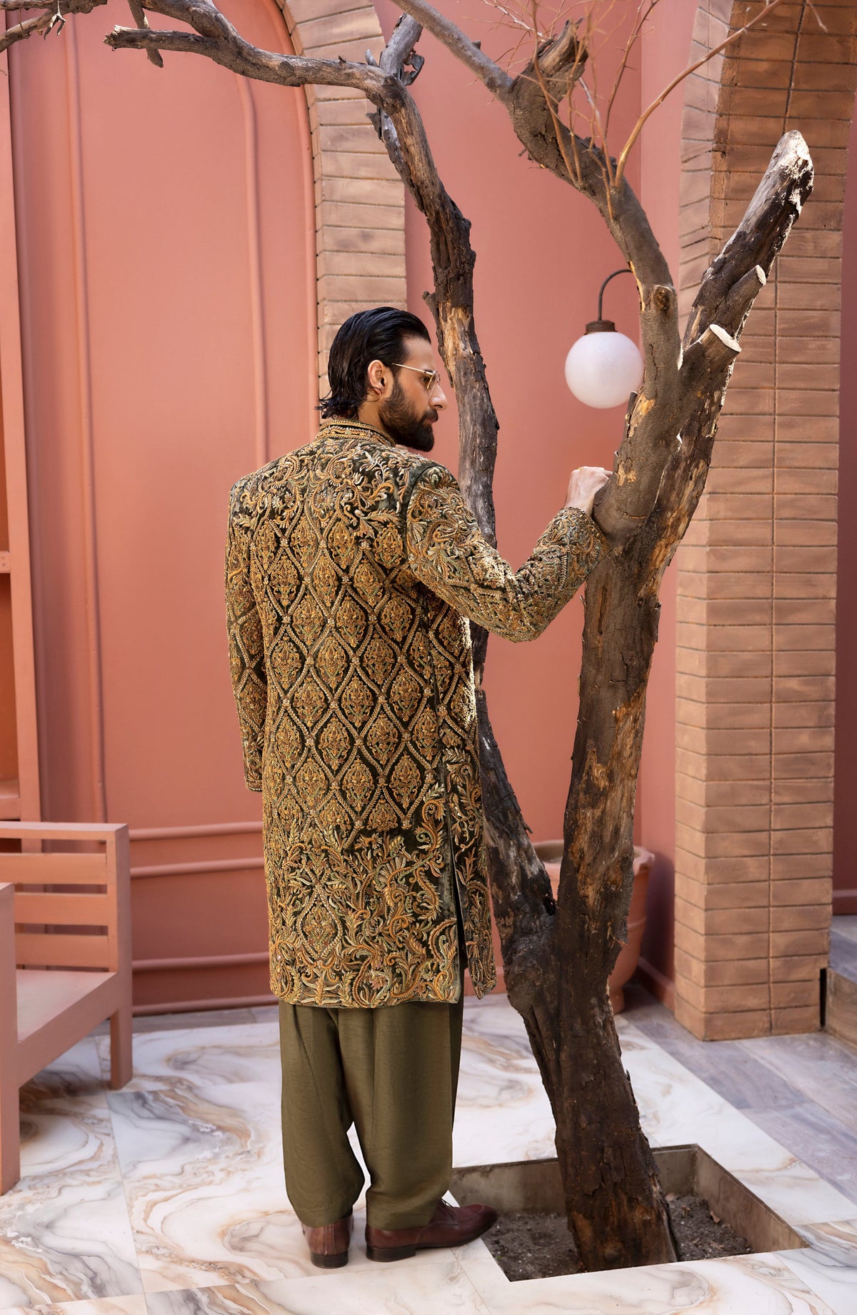A man wearing a gold and black heavily embroidered sherwani jacket with olive green trousers stands next to a decorative tree in a courtyard setting.