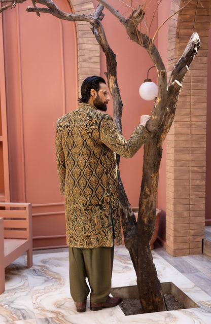 A man wearing a gold and black heavily embroidered sherwani jacket with olive green trousers stands next to a decorative tree in a courtyard setting.
