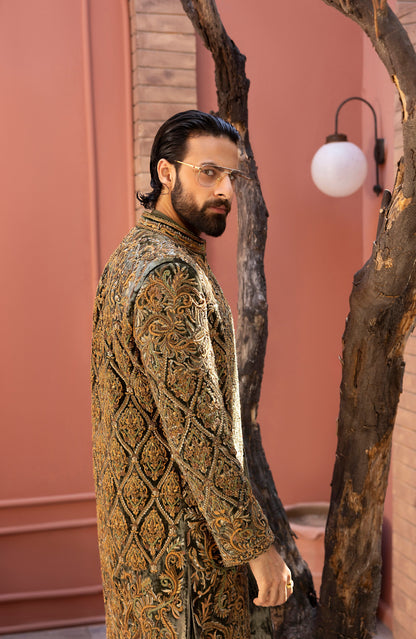 A man wearing a detailed, ornate gold and brown embroidered sherwani stands in profile against a dusty pink wall with a decorative tree trunk.