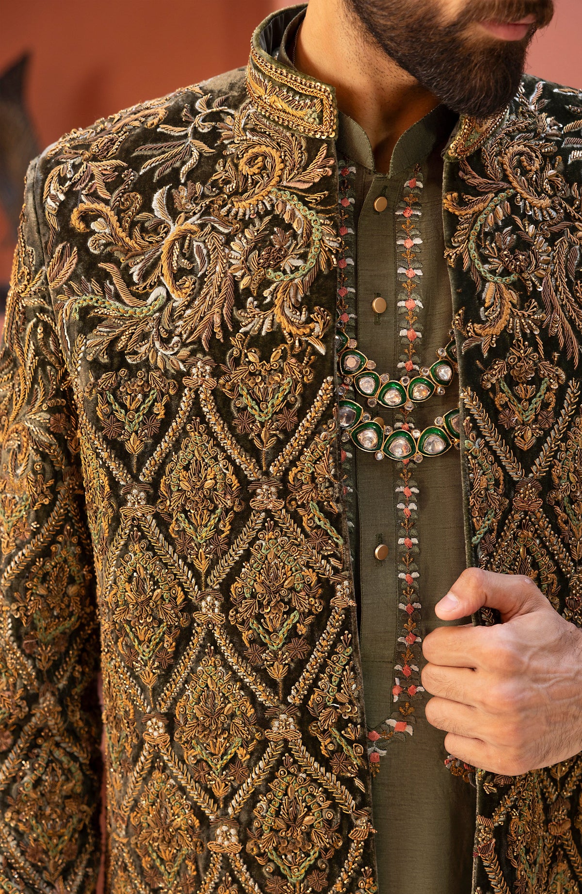 Close-up of a man wearing an ornate embroidered sherwani jacket over a matching olive green embroidered kurta. The jacket features intricate gold, brown, and green paisley and diamond-patterned embroidery.