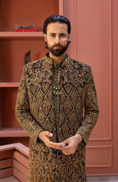 A man with a beard and glasses wearing a long-sleeved, mandarin-collar embroidered sherwani jacket in brown and gold, standing in front of a terracotta-colored paneled wall.
