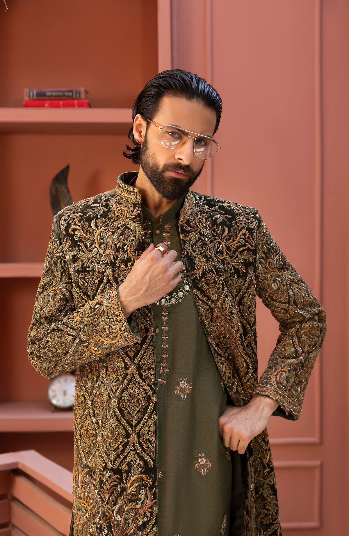 A man wearing a long-sleeved, gold and brown heavily embroidered sherwani jacket over an olive green kurta, standing in front of a dusty rose wooden bookshelf.