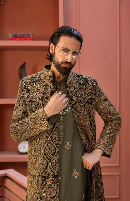 A man wearing a long-sleeved, gold and brown heavily embroidered sherwani jacket over an olive green kurta, standing in front of a dusty rose wooden bookshelf.