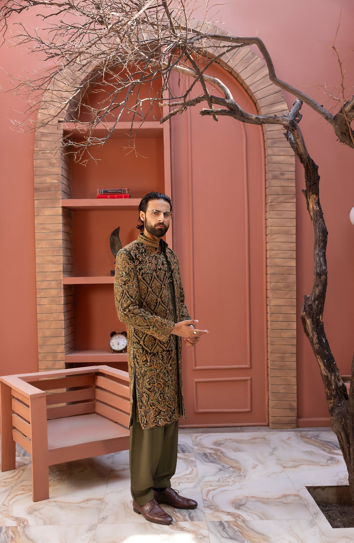 A man wearing a brown and gold patterned sherwani with olive green trousers stands in front of a terracotta-colored arched alcove with shelves and a bench.