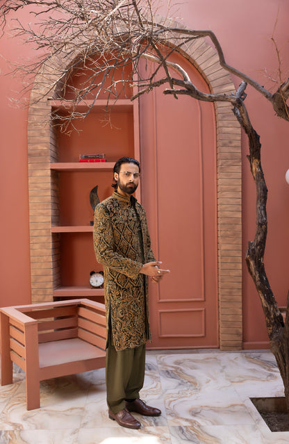 A man wearing a brown and gold patterned sherwani with olive green trousers stands in front of a terracotta-colored arched alcove with shelves and a bench.