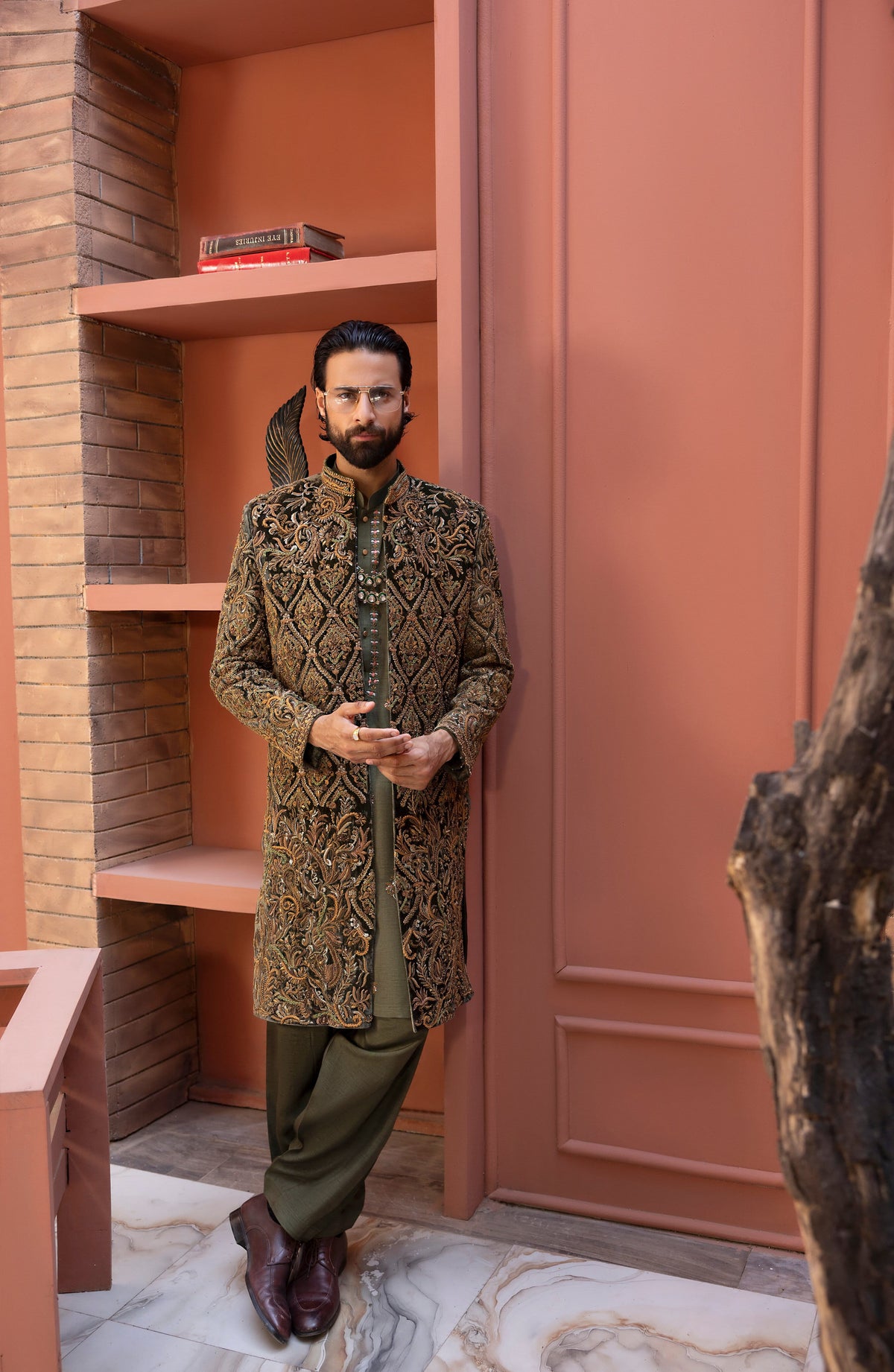 A man wearing a traditional long-sleeved embroidered sherwani in olive green and gold, paired with matching olive trousers and dark brown leather shoes, standing in a modern interior with terracotta-colored walls and built-in shelves.