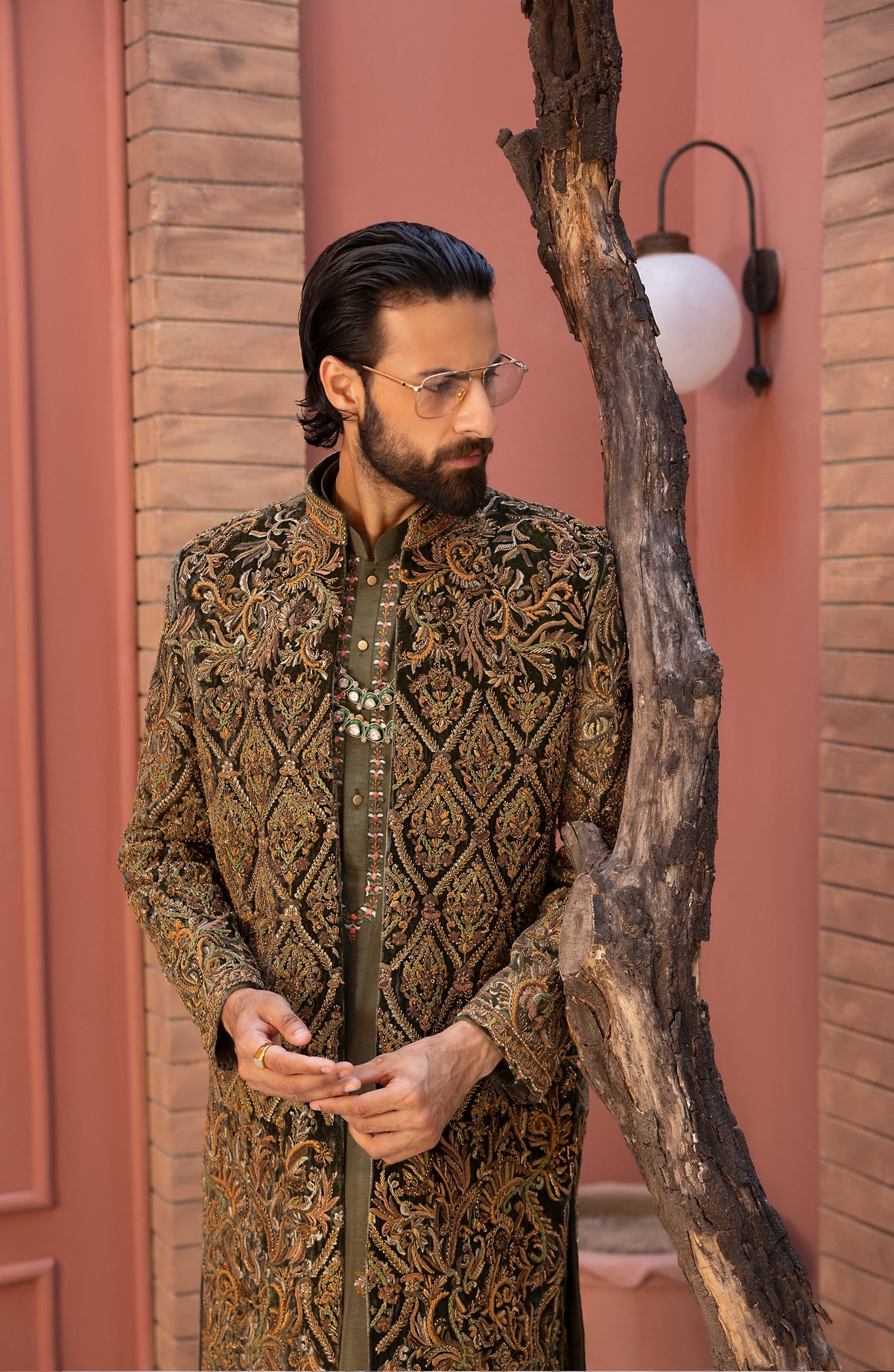 A man with a beard and glasses wearing a detailed gold and brown embroidered sherwani jacket, standing next to a rustic tree trunk in front of a pink brick wall.