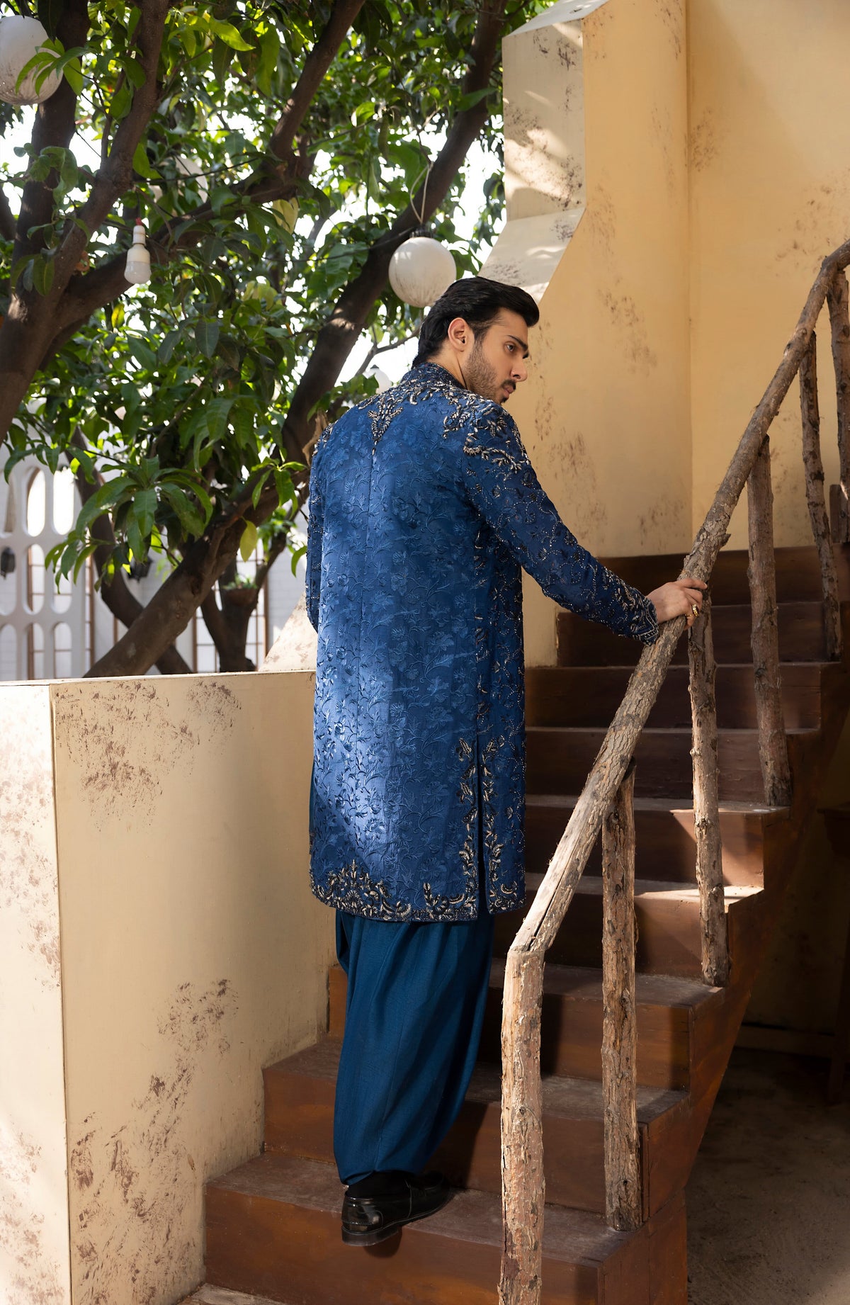 A man wearing a blue embroidered traditional kurta and trousers stands on wooden stairs holding a rustic handrail, with a tree and cream-colored building walls in the background.