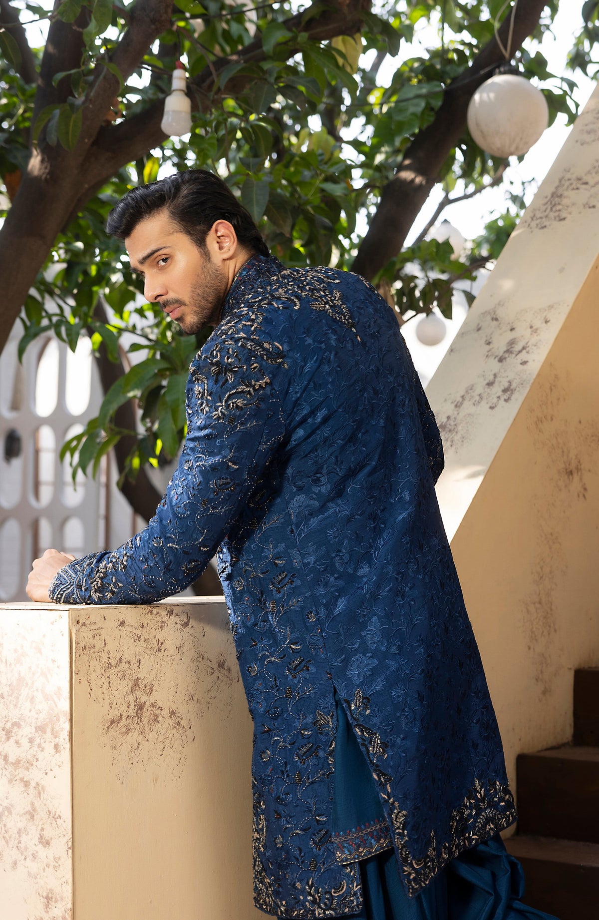 A man wearing a dark blue embroidered sherwani with gold detailing, leaning on a stone wall in an outdoor courtyard with trees and hanging lights.
