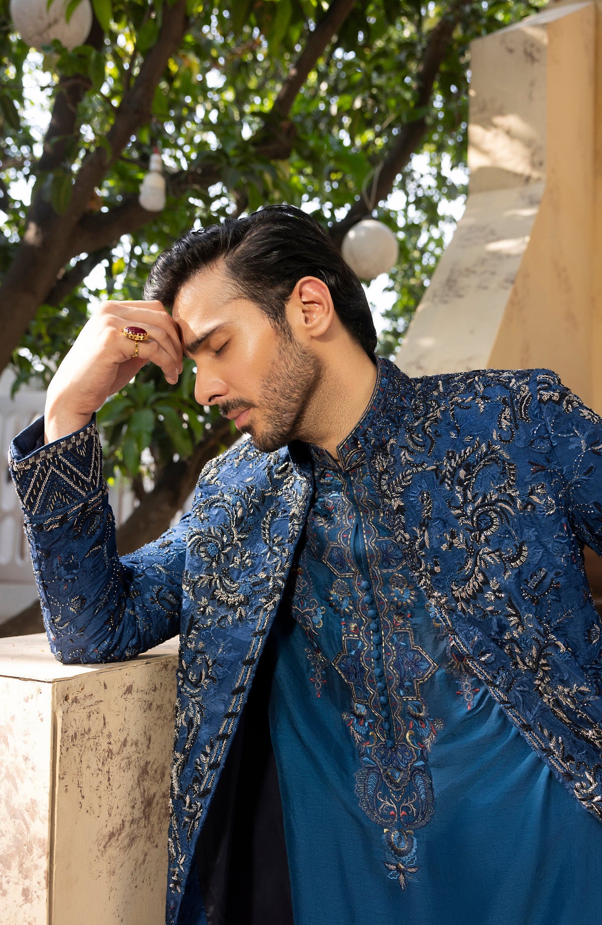 A man wearing a blue embroidered traditional kurta jacket with gold detailing, leaning on a stone wall outdoors under a tree.