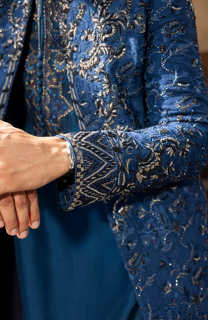 Close-up of a person wearing a rich royal blue velvet jacket with intricate silver and gold embroidery, paired with a matching blue kurta. The detailed sleeve cuff is shown with a silver bracelet on the wrist.