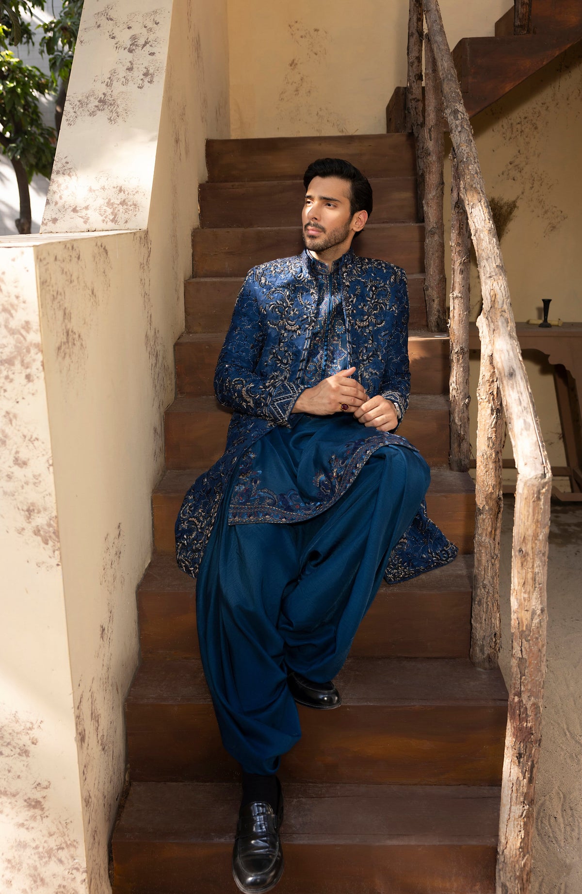 A man wearing a traditional Indian outfit with a blue patterned kurta and matching trousers sits on wooden stairs in an outdoor setting.